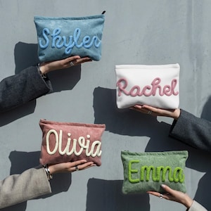 May include: Four corduroy pouches in blue, white, pink, and green, each with a name embroidered in a contrasting colour: Skyler, Rachel, Olivia, and Emma. The pouches are held up against a grey wall.