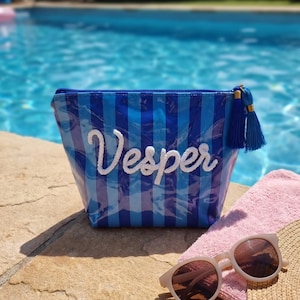 May include: A blue and light blue striped pouch with a glossy finish, featuring the white embroidered name Vesper on the front. The pouch includes a blue tassel attached to the zipper pull, providing a decorative accent to the accessory.