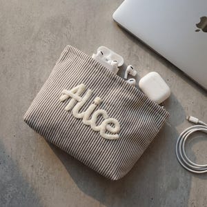 Op de afbeelding: Een beige en wit gestreept stoffen tasje met de naam Alice geborduurd in verhoogde witte schrijfletters. Het tasje is aan de bovenkant open en bevat witte draadloze oordopjes in hun oplaadcase en een witte oplaadkabel.