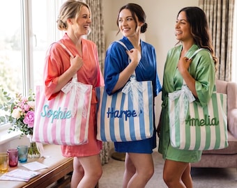 May include: Three tote bags with vertical stripes in pink, blue, and green, each featuring a white lace bow and embroidered names Sophie, Harper, and Amelia. The bags are carried by individuals wearing matching satin robes in corresponding colors.