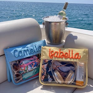 May include: Two rectangular clear pouches with wrist straps, one light blue with the name Camella embroidered in blue, and one yellow with the name Isabella embroidered in orange. Both contain various personal items like sunglasses and headphones.
