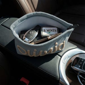 May include: A light blue corduroy car organizer with the name "David" embroidered in cream-colored yarn. The organizer contains a car key, a wallet, a pen, and a tin of B. Mints.