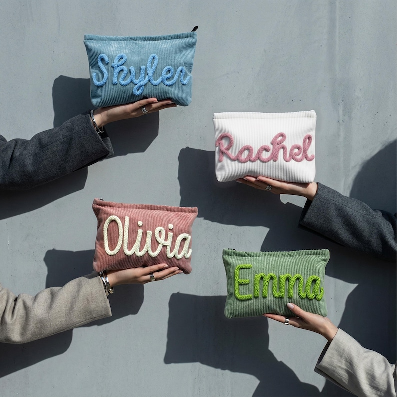 May include: Four corduroy pouches in blue, white, pink, and green, each with a name embroidered in a contrasting colour: Skyler, Rachel, Olivia, and Emma. The pouches are held up against a grey wall.