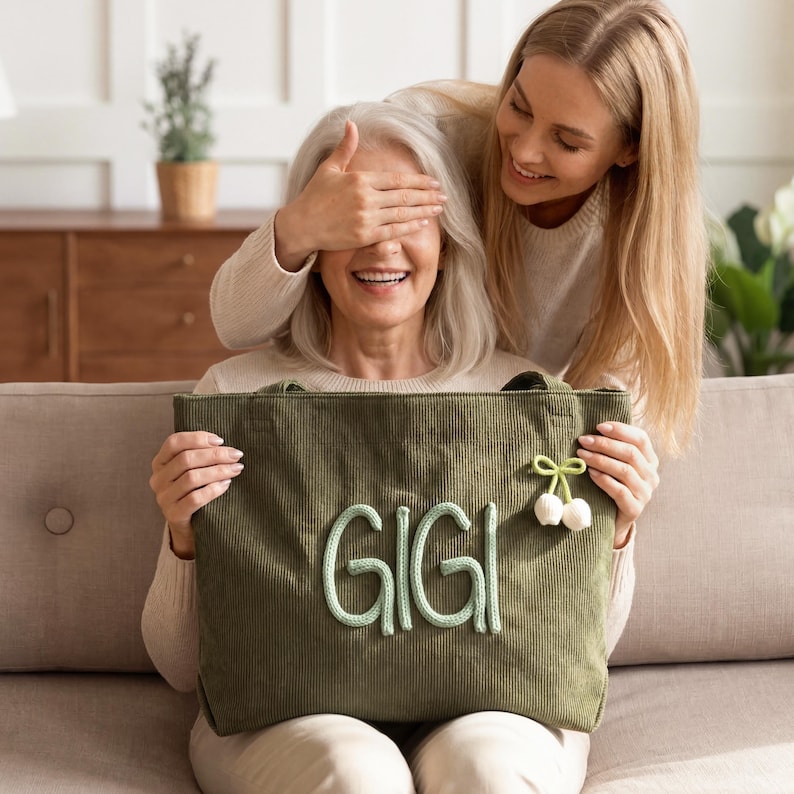 May include: An olive green corduroy tote bag featuring the name GIGI in raised, light green stitched lettering on the front. A small, decorative light green bow with two white dangling pom-poms is attached to the upper right side of the bag.