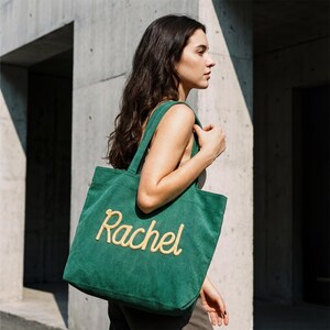 May include: Green corduroy tote bag with the name "Rachel" embroidered in a light yellow script. The bag has long straps and is carried over the shoulder. The tote is a solid green color.