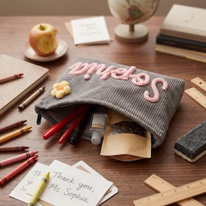 May include: A grey corduroy zipped pouch featuring the name Sophia embroidered in pink yarn and a small yellow crocheted flower appliqué. The pouch is open, revealing red pens, a clear plastic bottle, and a small brown paper bag inside.