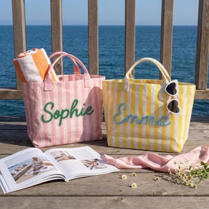 May include: Two striped canvas tote bags with handles. The pink and white bag features the name Sophie in green script, while the yellow and white bag features the name Emma in blue script. A pair of heart-shaped sunglasses hangs from the yellow bag.