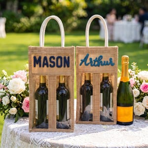 May include: Two rectangular burlap wine gift bags with white rope handles. One bag features the name MASON in dark blue block letters, and the other features the name Arthur in dark blue script. Each bag holds two wine bottles visible through clear windows.