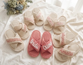May include: A collection of waffle-textured slippers with crisscross straps. Most are beige with pink embroidered names like 'bridesmaid', 'Sophia', 'Ana', 'Amelia', and 'Olivia'. Two slippers are pink with white embroidery reading 'bride' and 'Oliver'.