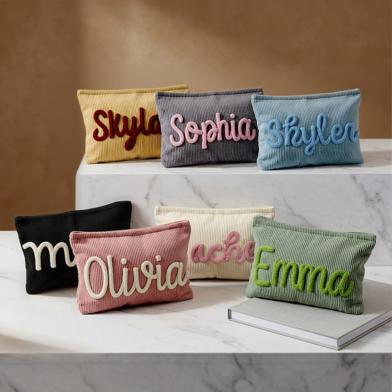 May include: A collection of rectangular corduroy pouches in various colors including yellow, gray, blue, black, pink, cream, and green. Each pouch features a raised, cursive-style fabric name appliqu&eacute; in a contrasting color, reading Skyla, Sophia, Skyler, m, Olivia, rache, and Emma.