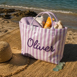 May include: A tote bag featuring vertical lavender and white stripes with the name Oliver embroidered in dark purple script on the front. The bag contains a folded purple towel, a beige fabric item, and an orange sunscreen bottle.