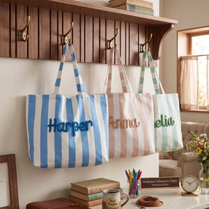Peut inclure: Trois sacs fourre-tout en toile &agrave; rayures verticales bleues, roses et vertes, chacun arborant un nom brod&eacute; en relief sur le devant. Les sacs sont suspendus &agrave; des crochets en laiton sur un support en bois, affichant les noms Harper, Anna et Amelia.