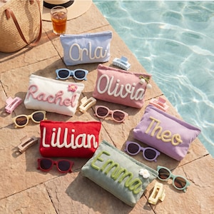 May include: A collection of personalized cosmetic bags in various colors, each embroidered with a name in a cursive font. Sunglasses and hair clips are arranged around the bags on a stone surface near a pool.