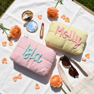 Puede incluir: Un estuche acolchado rosa con el nombre Isla en letras de cuerda azules y un estuche acolchado amarillo con el nombre Molly en letras de cuerda rosas, junto a un espejo compacto dorado, un pintalabios, gafas de sol y flores ranúnculos naranjas.
