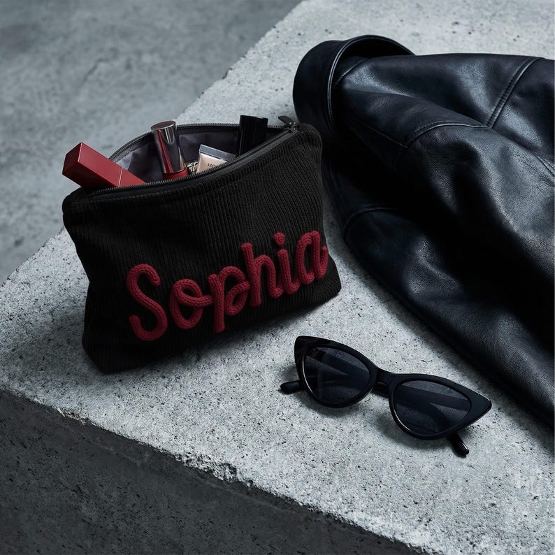 May include: A dark brown ribbed knit zippered pouch with the name Sophia embroidered in red script lettering. The pouch contains various cosmetic items, including a red lipstick tube. Beside it are black cat-eye sunglasses and a black leather jacket.