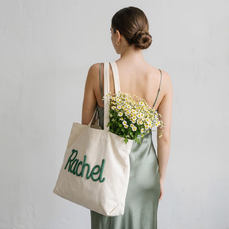 May include: Cream-colored corduroy tote bag with the name "Rachel" embroidered in green script. The bag is filled with a bouquet of white and yellow flowers. The person is wearing a sage green dress.