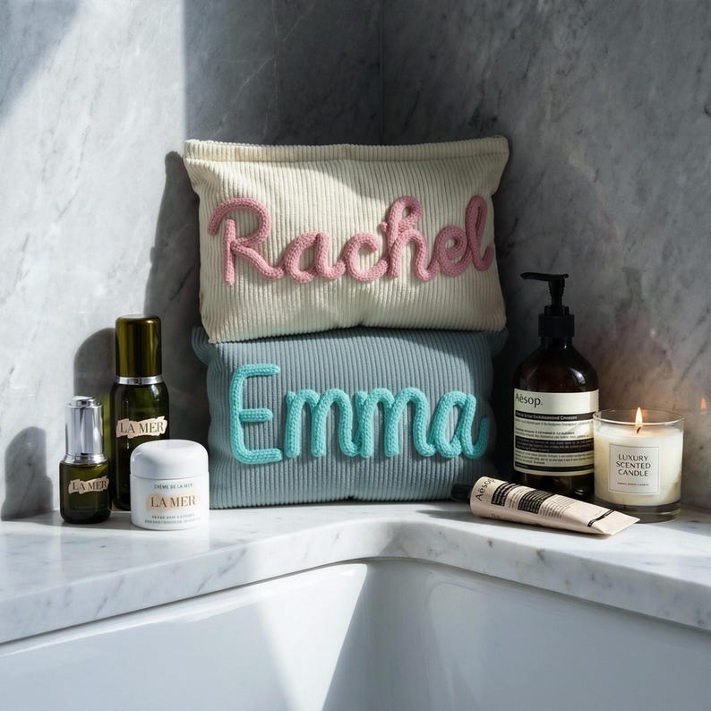 May include: Two rectangular corduroy pouches stacked vertically. The top cream pouch features the name Rachel in pink script, while the bottom sage green pouch displays the name Emma in teal script. Various skincare bottles and a candle surround them.