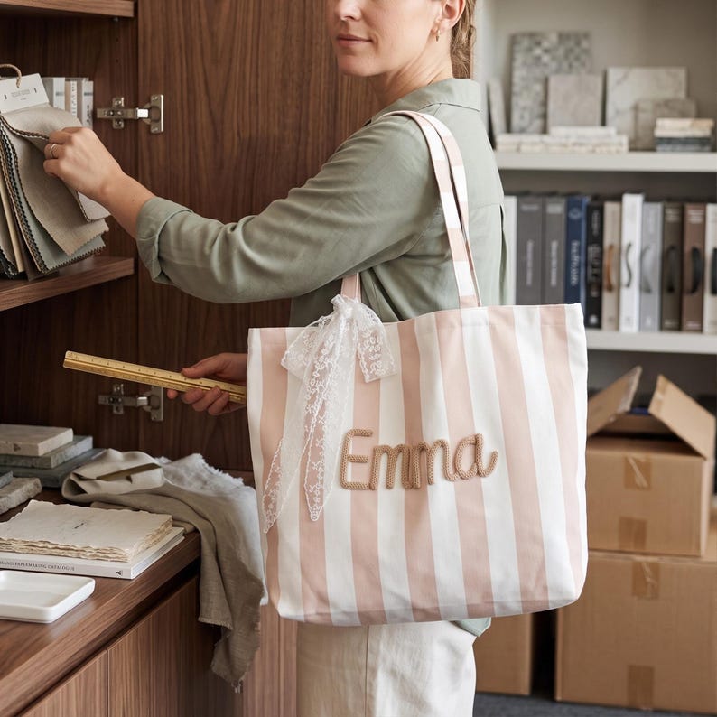 Peut inclure: Un sac fourre-tout &agrave; rayures verticales rose clair et blanches avec le pr&eacute;nom Emma brod&eacute; en fil beige sur le devant. Le sac comprend deux bandouli&egrave;res et un ruban en dentelle blanche nou&eacute; en n&oelig;ud sur le c&ocirc;t&eacute; de l'attache de la poign&eacute;e.