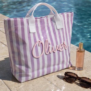 May include: A tote bag featuring vertical lavender and white stripes with white fabric handles. The name Oliver is written across the front in raised pink rope lettering. A clear glass bottle with amber liquid and tortoise shell sunglasses are nearby.