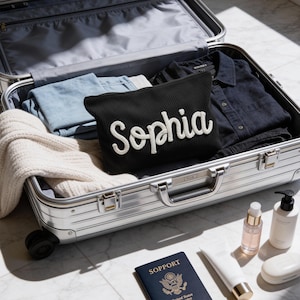May include: A black ribbed zippered pouch with the white embroidered name Sophia, placed inside an open silver metallic suitcase containing folded light blue and black clothing, a cream sweater, a passport, and various white toiletry bottles.