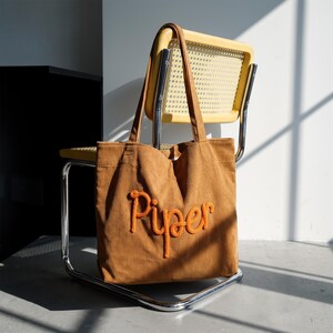 May include: A brown corduroy tote bag with the name "Piper" embroidered in orange. The bag has long handles and is resting on a yellow chair with a woven back. The background is a white wall and a black cabinet.