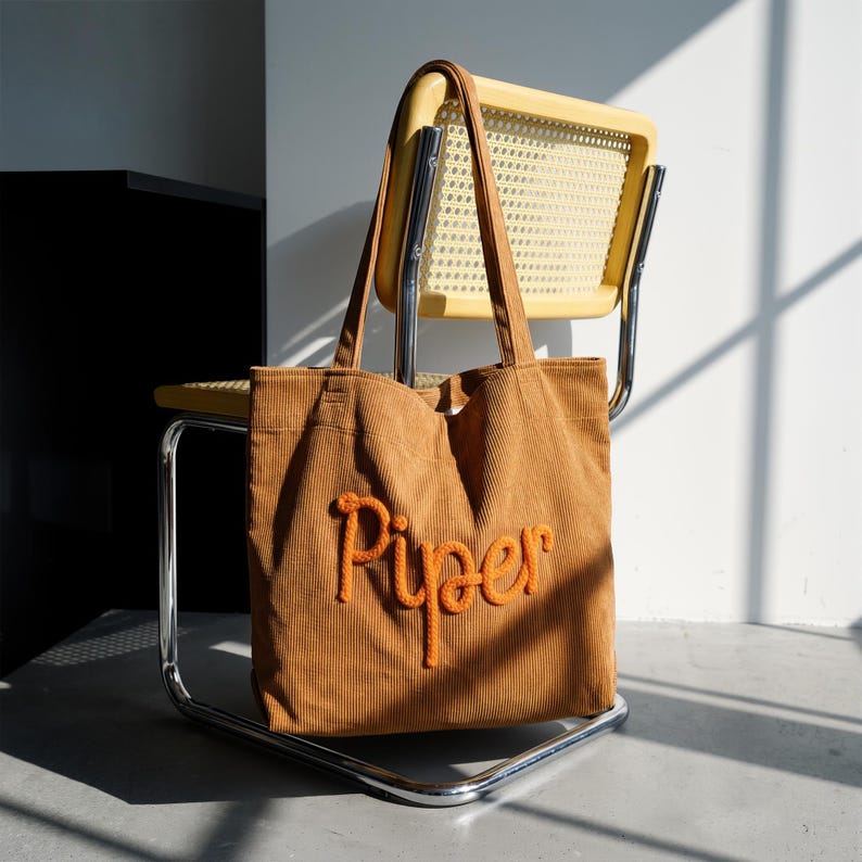 May include: A brown corduroy tote bag with the name "Piper" embroidered in orange. The bag has long handles and is resting on a yellow chair with a woven back. The background is a white wall and a black cabinet.