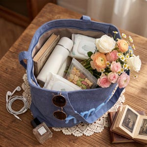 May include: A blue corduroy tote bag filled with a white thermos, a book titled Moments Remembered, two packs of wet wipes, a clear pouch of colorful snacks, and a bouquet of white, pink, and orange flowers. Round sunglasses hang from the bag strap.
