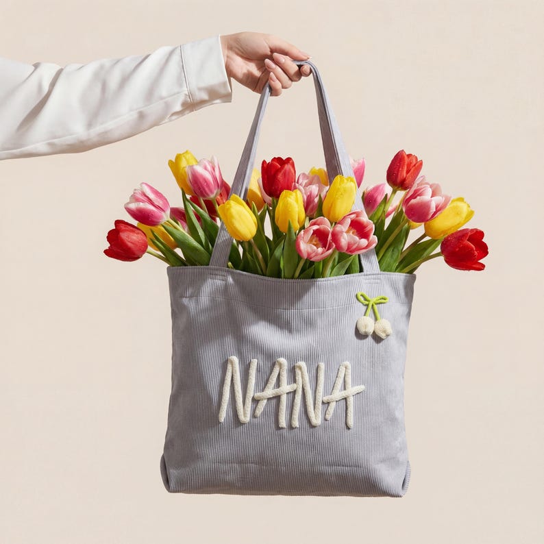 May include: A light blue corduroy tote bag featuring the word NANA in white embroidered lettering on the front. A small green and white cherry-shaped patch is attached to the upper right corner. The bag is filled with red, yellow, and pink tulips.