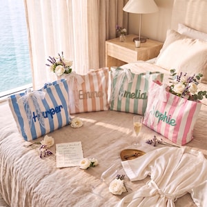 Peut inclure: Quatre sacs fourre-tout ray&eacute;s en bleu, p&ecirc;che, vert et rose, chacun orn&eacute; d'un n&oelig;ud en dentelle blanche et d'un pr&eacute;nom brod&eacute; dans une couleur assortie. Les sacs sont dispos&eacute;s sur un lit avec des fleurs blanches, une carte et une robe blanche.