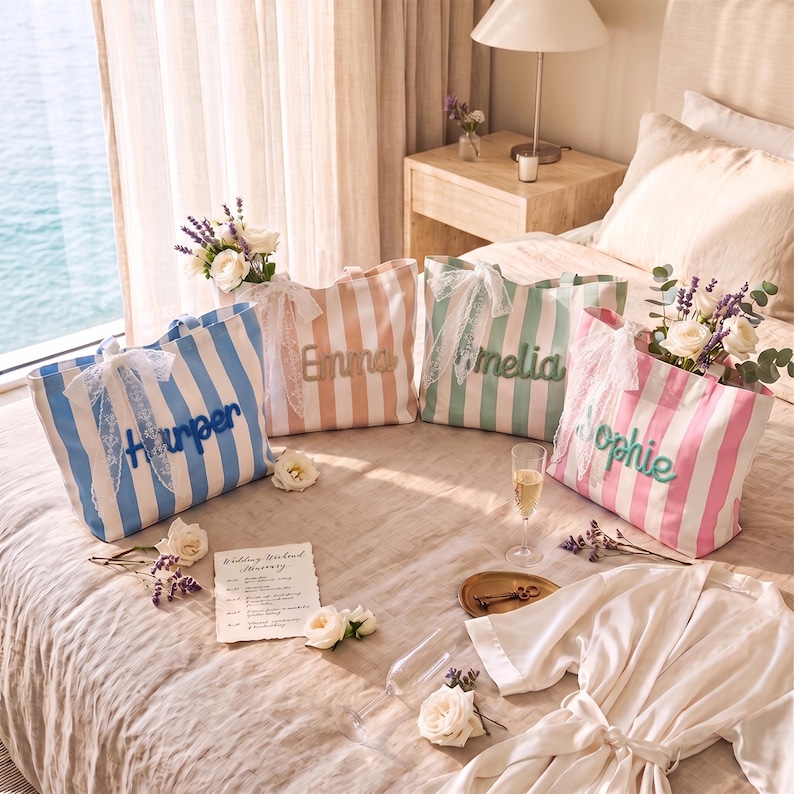 Peut inclure: Quatre sacs fourre-tout ray&eacute;s en bleu, p&ecirc;che, vert et rose, chacun orn&eacute; d'un n&oelig;ud en dentelle blanche et d'un pr&eacute;nom brod&eacute; dans une couleur assortie. Les sacs sont dispos&eacute;s sur un lit avec des fleurs blanches, une carte et une robe blanche.