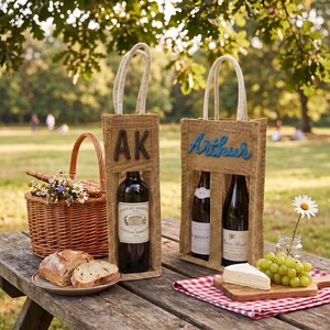 May include: Two tan burlap wine bags with rope handles. One bag holds a single wine bottle and features the black embroidered letters AK. The other bag holds two wine bottles and features the blue embroidered name Arthur on the front.
