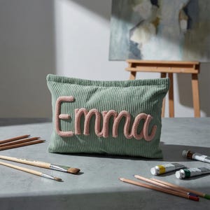 May include: A rectangular sage green corduroy pouch featuring the name Emma in raised, soft pink knitted lettering centered on the front. The pouch has a textured ribbed fabric surface and is positioned among various wooden pencils and paint tubes.