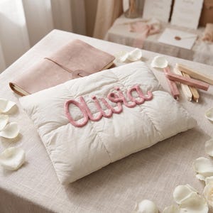 May include: A rectangular off-white quilted pillow featuring the name Olivia written in raised, pink knitted yarn script across the center. The pillow has a soft, padded texture with visible stitching lines forming a grid pattern on the fabric surface.