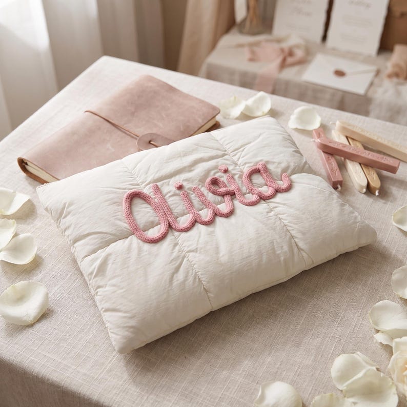 May include: A rectangular off-white quilted pillow featuring the name Olivia written in raised, pink knitted yarn script across the center. The pillow has a soft, padded texture with visible stitching lines forming a grid pattern on the fabric surface.