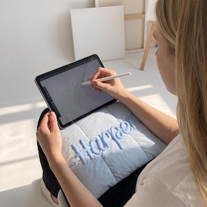 May include: A light blue quilted pillow featuring the name Harper embroidered in a matching light blue yarn script font. The pillow is resting on a lap while a tablet with a stylus is being used to draw on the screen above the pillow.