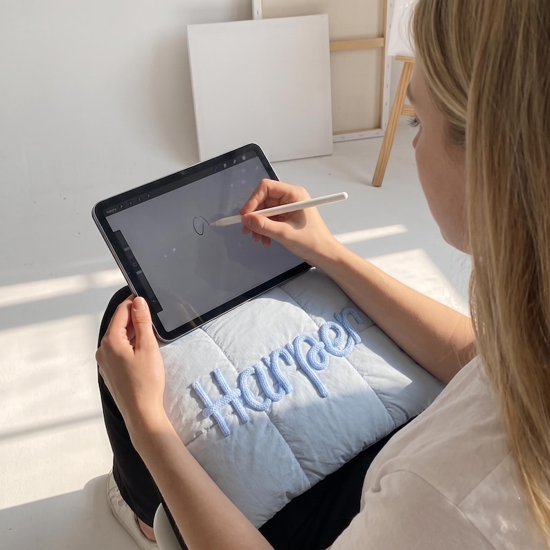 May include: A light blue quilted pillow featuring the name Harper embroidered in a matching light blue yarn script font. The pillow is resting on a lap while a tablet with a stylus is being used to draw on the screen above the pillow.