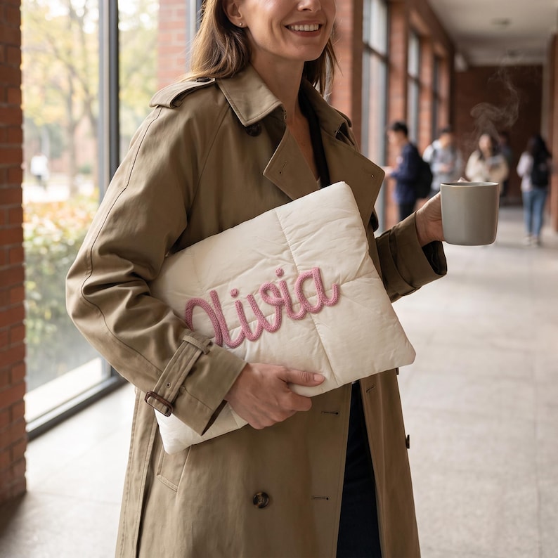 May include: A rectangular, off-white quilted laptop sleeve featuring the name Olivia embroidered in raised, dusty pink cursive lettering across the front. The sleeve has a soft, padded texture and a clean, minimalist design for carrying electronics.