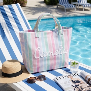 May include: A tote bag featuring vertical pastel stripes in pink, blue, and green with the name Camelia embroidered in white script on the front. The bag has two matching striped handles and a structured rectangular shape for carrying items.
