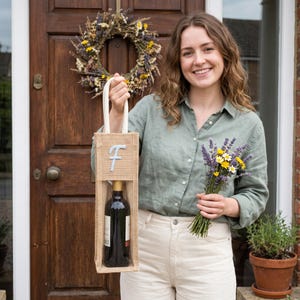 May include: A tan burlap wine gift bag featuring a white fabric handle and a light blue embroidered letter F on the front. The bag has a rectangular cutout window displaying a dark glass wine bottle with a gold foil neck label inside.