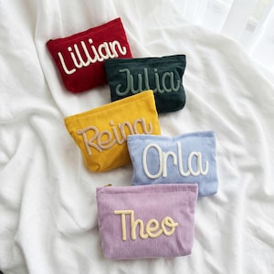 May include: Five rectangular corduroy zipper pouches in red, dark green, yellow, light blue, and lavender. Each pouch features a name embroidered in a raised, cursive font: Lillian, Julia, Reina, Orla, and Theo, with matching or contrasting thread.