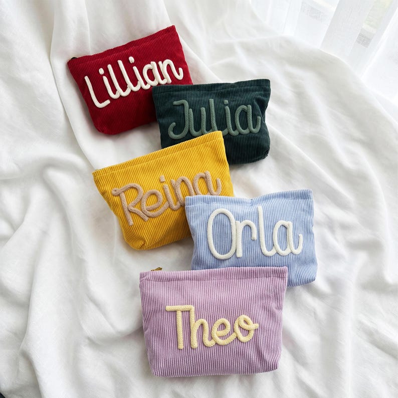 May include: Five rectangular corduroy zipper pouches in red, dark green, yellow, light blue, and lavender. Each pouch features a name embroidered in a raised, cursive font: Lillian, Julia, Reina, Orla, and Theo, with matching or contrasting thread.