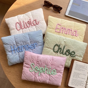 Könnte beinhalten: Fünf rechteckige, gesteppte Taschen in Pastellfarben, jede mit einem in erhabenem Garn gestickten Namen: Olivia in Pink, Emma in Pink, Harper in Blau, Chloe in Dunkelgrün und Sophie in Hellgrün. Die Taschen sind zusammen angeordnet.