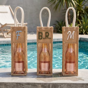 May include: Three vertical burlap wine gift bags with white rope handles and clear plastic windows. Each bag features a unique embroidered letter or monogram: a light blue F, dark green B.R., and white M, each containing a glass bottle of rose wine.