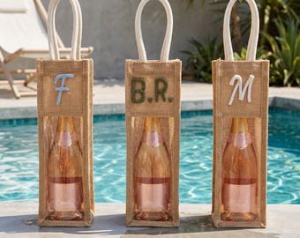 May include: Three vertical burlap wine gift bags with white rope handles and clear plastic windows. Each bag features a unique embroidered letter or monogram: a light blue F, dark green B.R., and white M, each containing a glass bottle of rose wine.
