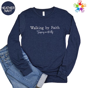 A navy blue long-sleeved shirt with the text "Walking by Faith" printed on it, along with the phrase "Tripping constantly" below it. The shirt is laid out on a white background, with a pair of blue jeans visible underneath.