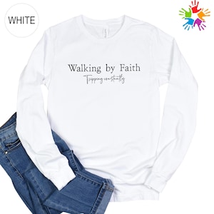 A white long-sleeved t-shirt with the text "Walking by Faith" printed on it, along with the phrase "Tripping constantly" below it. The t-shirt is placed on top of a pair of blue jeans.