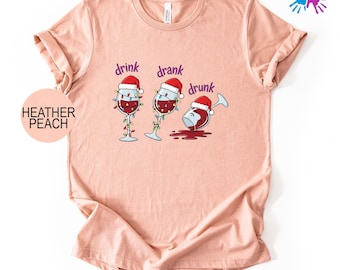 May include: Heather peach-colored t-shirt with a humorous graphic of three wine glasses wearing Santa hats. The glasses are strung with Christmas lights, with the words "drink, drank, drunk" above them.