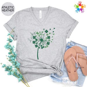 May include: A light gray V-neck t-shirt with a green dandelion design made of shamrocks and the word "lucky" in script. The shirt is paired with denim shorts and pink sandals. The shirt is labeled "Athletic Heather".
