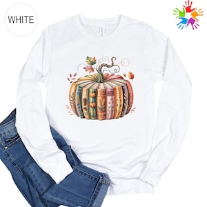 May include: A white long-sleeve shirt featuring a graphic of a pumpkin constructed from a circular arrangement of colorful books. The books are decorated with illustrations of wheat, acorns, and flowers, topped with a stem and autumn leaves.