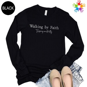 a black long-sleeved shirt with the text "Walking by Faith" printed on it, along with the phrase "Tripping constantly" below it. The shirt is laid out on a white background, and there are beige shoes visible underneath.